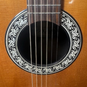 May include: Close-up of an acoustic guitar. The guitar features a light brown wooden body with a black soundhole. The soundhole is surrounded by a white and black decorative design. Six strings are visible.