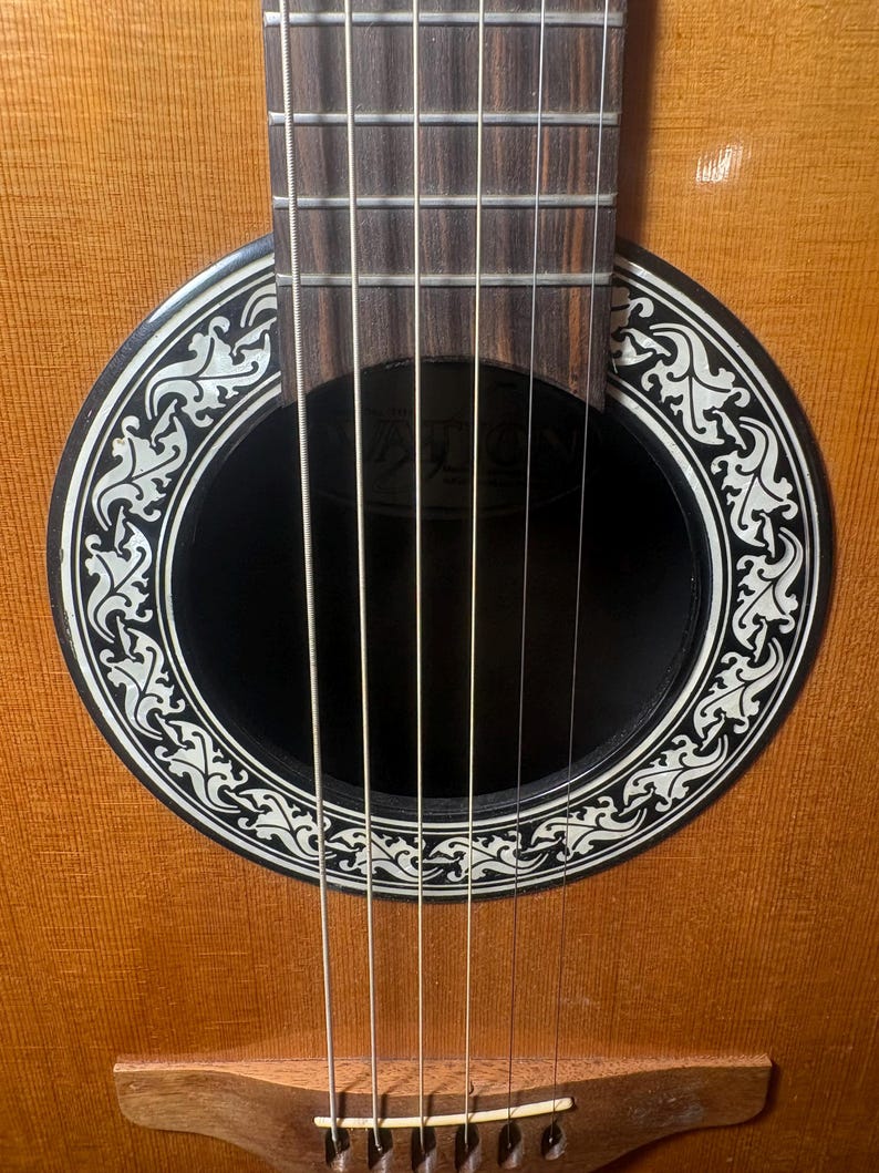 May include: Close-up of an acoustic guitar. The guitar features a light brown wooden body with a black soundhole. The soundhole is surrounded by a white and black decorative design. Six strings are visible.
