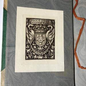 Puede incluir: Libro abierto con una ilustraci&oacute;n impresa. La ilustraci&oacute;n es un escudo de armas detallado en blanco y negro con elementos florales y de aves, y el texto "FRANCES EVELYN COUNTESS OF WARWICK". El libro tiene una columna vertebral verde y p&aacute;ginas grises veteadas.