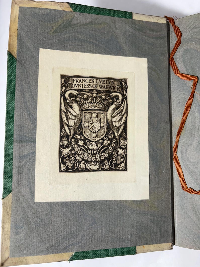 Puede incluir: Libro abierto con una ilustraci&oacute;n impresa. La ilustraci&oacute;n es un escudo de armas detallado en blanco y negro con elementos florales y de aves, y el texto "FRANCES EVELYN COUNTESS OF WARWICK". El libro tiene una columna vertebral verde y p&aacute;ginas grises veteadas.
