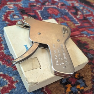 May include: A metallic Hesse-Majestic Lockaid tool with a trigger mechanism and a small wheel, resting on a white box. The tool is engraved with the text Hesse-Majestic Lockaid and Pat No. 1997362. The device has a silver-toned finish and a pistol-like shape.