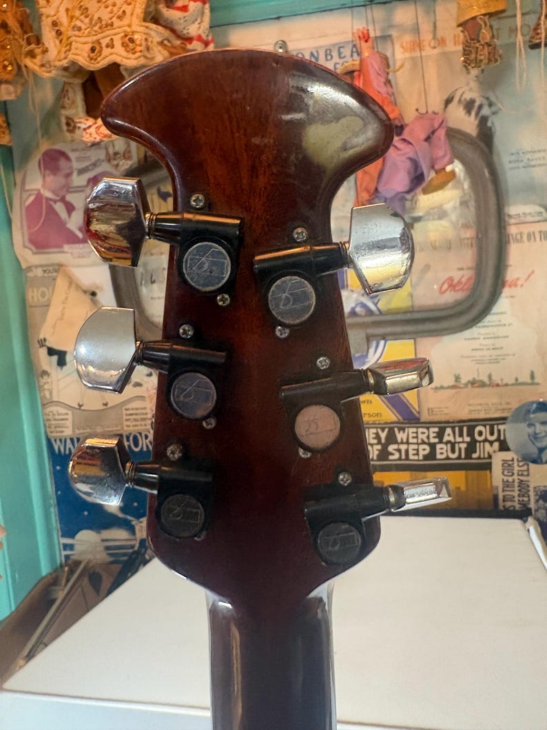 May include: Close-up of the headstock of a brown acoustic guitar. The headstock features six chrome tuning pegs and black tuning machines. The wood has a glossy finish with some wear and tear.
