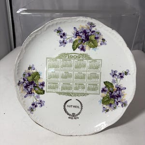 May include: White ceramic plate with a scalloped edge, featuring a 1909 calendar design. The plate is decorated with purple floral clusters and the text "GUT HEIL" in a decorative wreath.