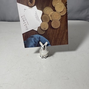 Puede incluir: Una pequeña figura de conejo plateado con base blanca. La figura está frente a una superficie de madera con monedas doradas y un papel que dice "Collect". El conejo tiene orejas erguidas y una cara detallada.