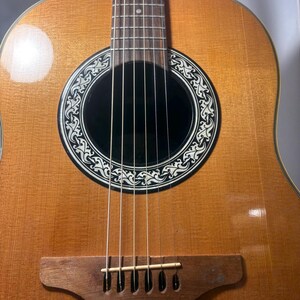 May include: Acoustic guitar with a light brown wooden body and a dark sound hole. The sound hole is surrounded by a decorative black and white design. The guitar has six strings and a wooden bridge.