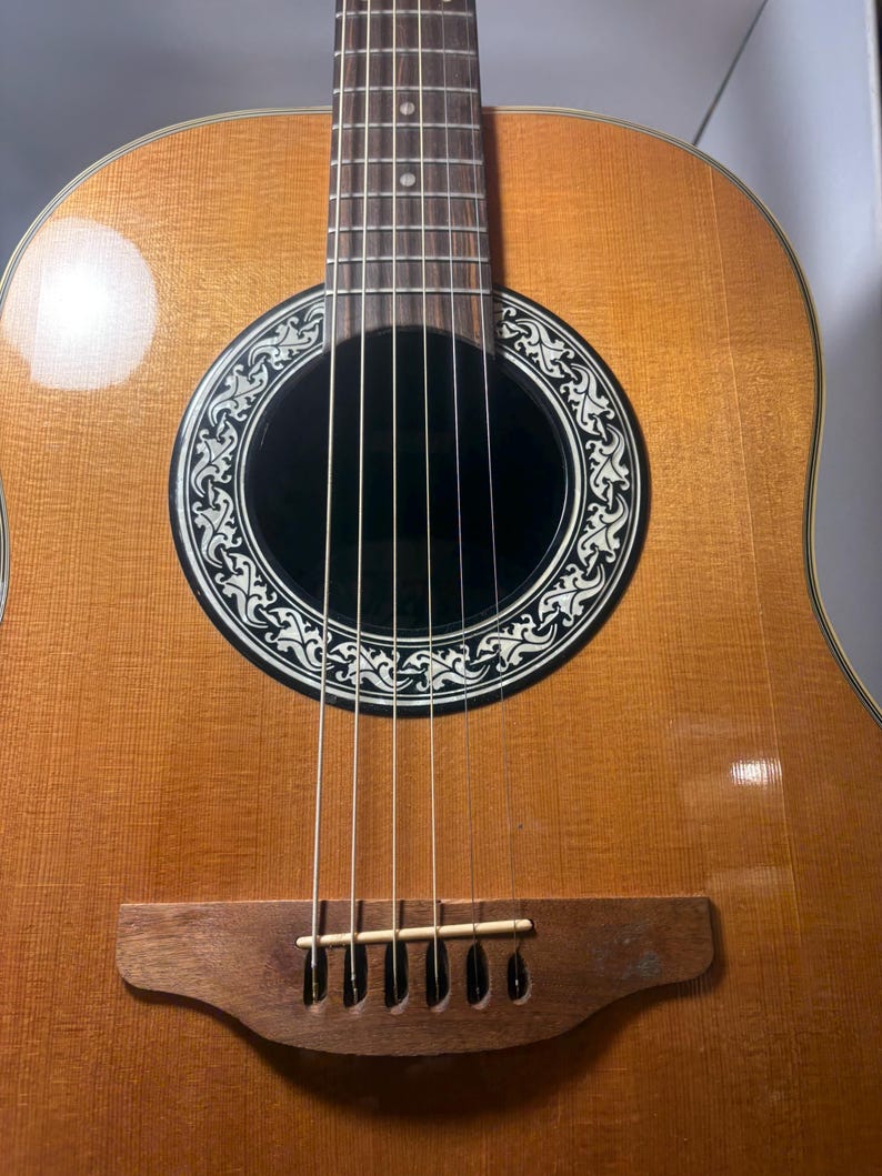May include: Acoustic guitar with a light brown wooden body and a dark sound hole. The sound hole is surrounded by a decorative black and white design. The guitar has six strings and a wooden bridge.