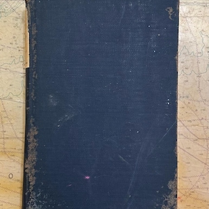 Könnte beinhalten: Ein rechteckiges Hardcover-Buch mit einem dunkelblauen, strukturierten Stoffeinband. Die Buchrücken-Kante weist deutliche Abnutzungserscheinungen mit ausgefransten Rändern und freiliegendem hellem Material auf. Die Oberfläche zeigt leichte Kratzer.