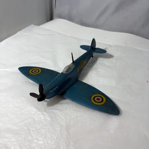 May include: A teal-colored model airplane with black propellers and a clear cockpit. The wings feature yellow and red circular markings. The text "BT OK" is visible on the fuselage. This vintage toy airplane is a collectible item.