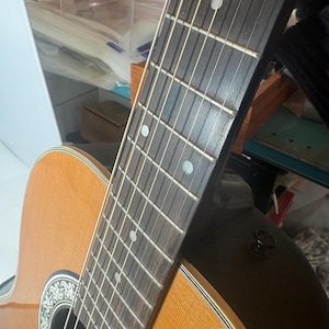 May include: Close-up of an acoustic guitar. The guitar features a light brown wooden body with a dark brown neck and fretboard. The strings are visible, and the sound hole has a decorative design. The guitar is angled, showing its side and neck.