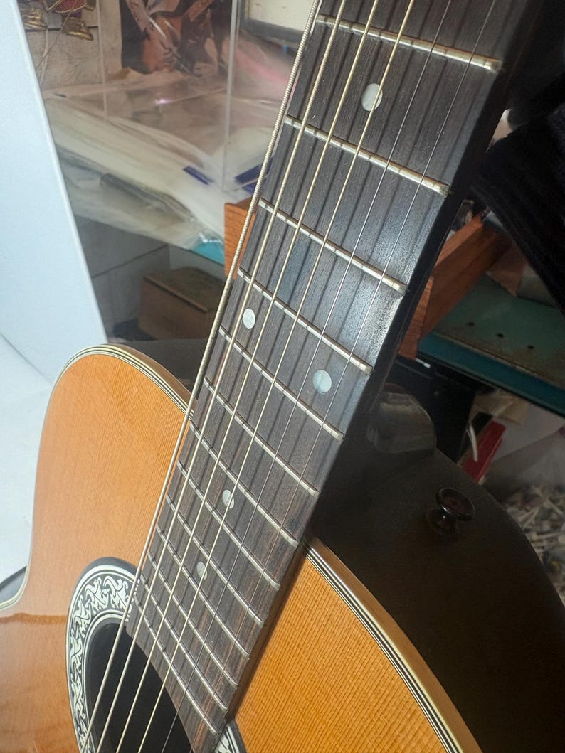 May include: Close-up of an acoustic guitar. The guitar features a light brown wooden body with a dark brown neck and fretboard. The strings are visible, and the sound hole has a decorative design. The guitar is angled, showing its side and neck.