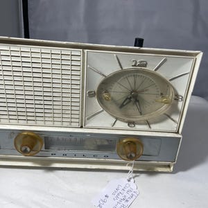 Peut inclure: Radio-réveil Philco vintage avec un cadran blanc et argenté. La radio a un haut-parleur à gauche et une horloge avec un cadran transparent à droite. Il comporte deux boutons ambrés et la marque "PHILCO" est visible.