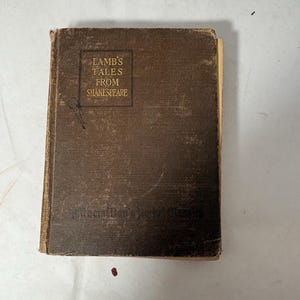 Puede incluir: Un libro antiguo de tapa dura marrón, titulado "Lamb's Tales From Shakespeare". La portada presenta letras doradas dentro de un marco rectangular. Los bordes del libro están desgastados, lo que sugiere una edición vintage.