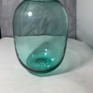 May include: A teal-coloured glass vase with a rounded body and a wide opening. The vase has a smooth surface and a slightly textured base. The glass is thick and appears hand-blown. The vase is empty and sits on a white surface.