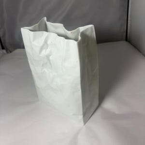 Puede incluir: Un florero de cer&aacute;mica blanca con forma de bolsa de papel arrugada. El objeto presenta una base rectangular, lados verticales y un borde superior irregular y plegado que imita la apariencia de una bolsa de papel aplastada con acabado mate.