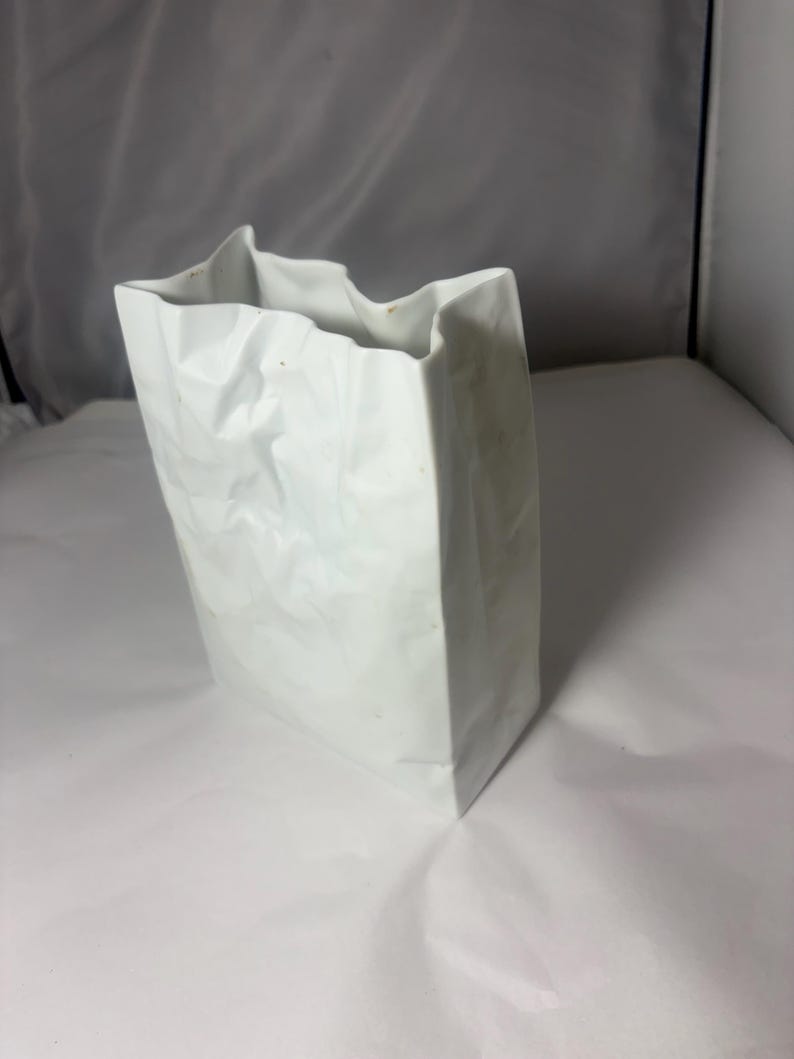 Puede incluir: Un florero de cer&aacute;mica blanca con forma de bolsa de papel arrugada. El objeto presenta una base rectangular, lados verticales y un borde superior irregular y plegado que imita la apariencia de una bolsa de papel aplastada con acabado mate.