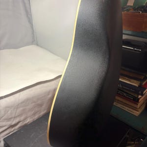 May include: Acoustic guitar with a black textured back and sides. The edge of the guitar has a light wood trim. The guitar is resting on a black surface.