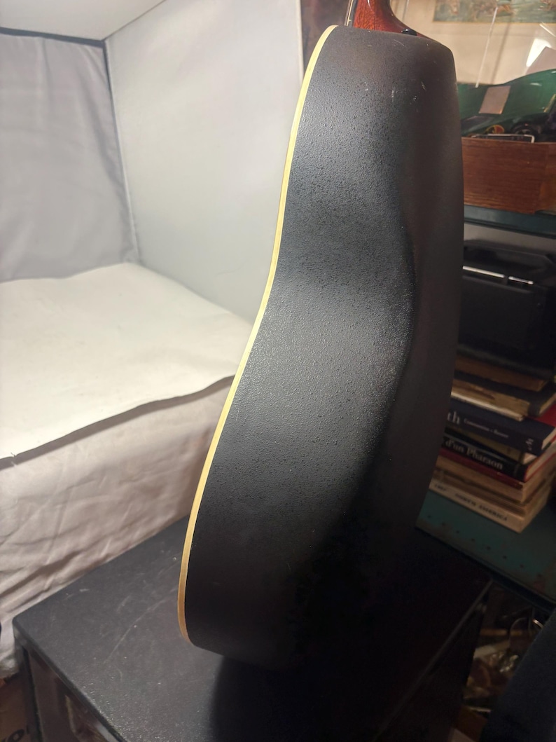 May include: Acoustic guitar with a black textured back and sides. The edge of the guitar has a light wood trim. The guitar is resting on a black surface.