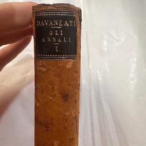May include: The spine of an antique book with a worn, light brown textured cover. A black rectangular label is affixed to the top, featuring gold-colored text that reads DAVANZATI, GLI ANNALI, I, enclosed within a decorative border.