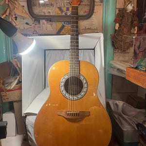 May include: Acoustic-electric guitar with a light brown wooden body and a round sound hole with a decorative rosette. The guitar has a wooden neck and headstock, with six strings and a bridge. The guitar is resting on a white surface.
