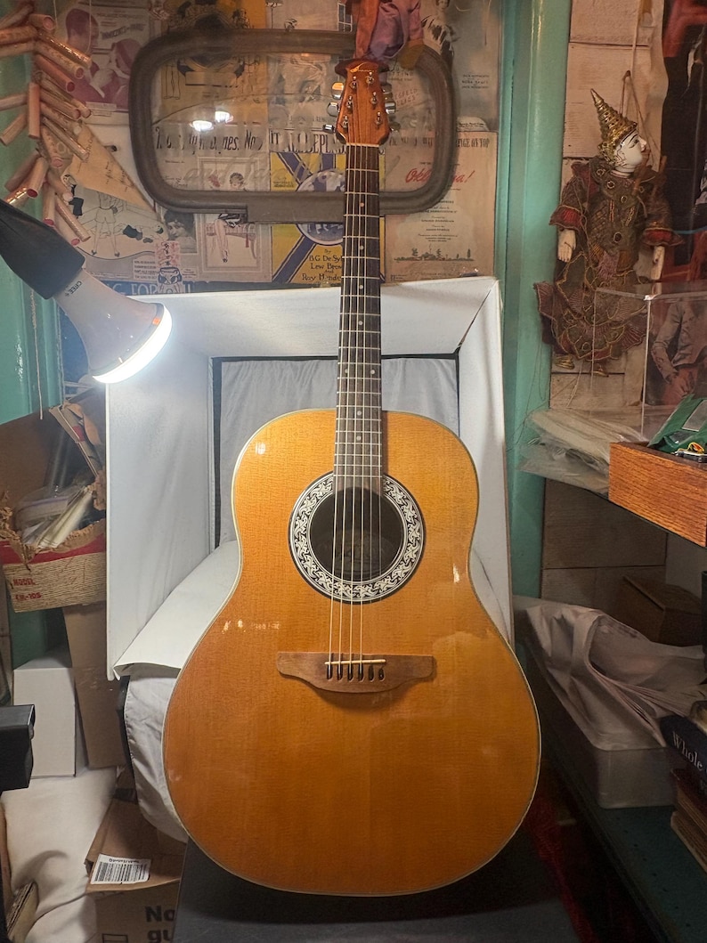 May include: Acoustic-electric guitar with a light brown wooden body and a round sound hole with a decorative rosette. The guitar has a wooden neck and headstock, with six strings and a bridge. The guitar is resting on a white surface.