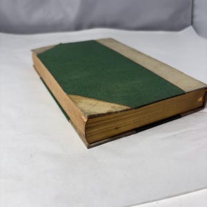 Puede incluir: Un libro antiguo con una cubierta verde y bordes beige. El lomo del libro es verde y los bordes de las p&aacute;ginas est&aacute;n envejecidos. La cubierta tiene un aspecto texturizado y las esquinas muestran desgaste. El libro es probablemente una antig&uuml;edad o un objeto de colecci&oacute;n.