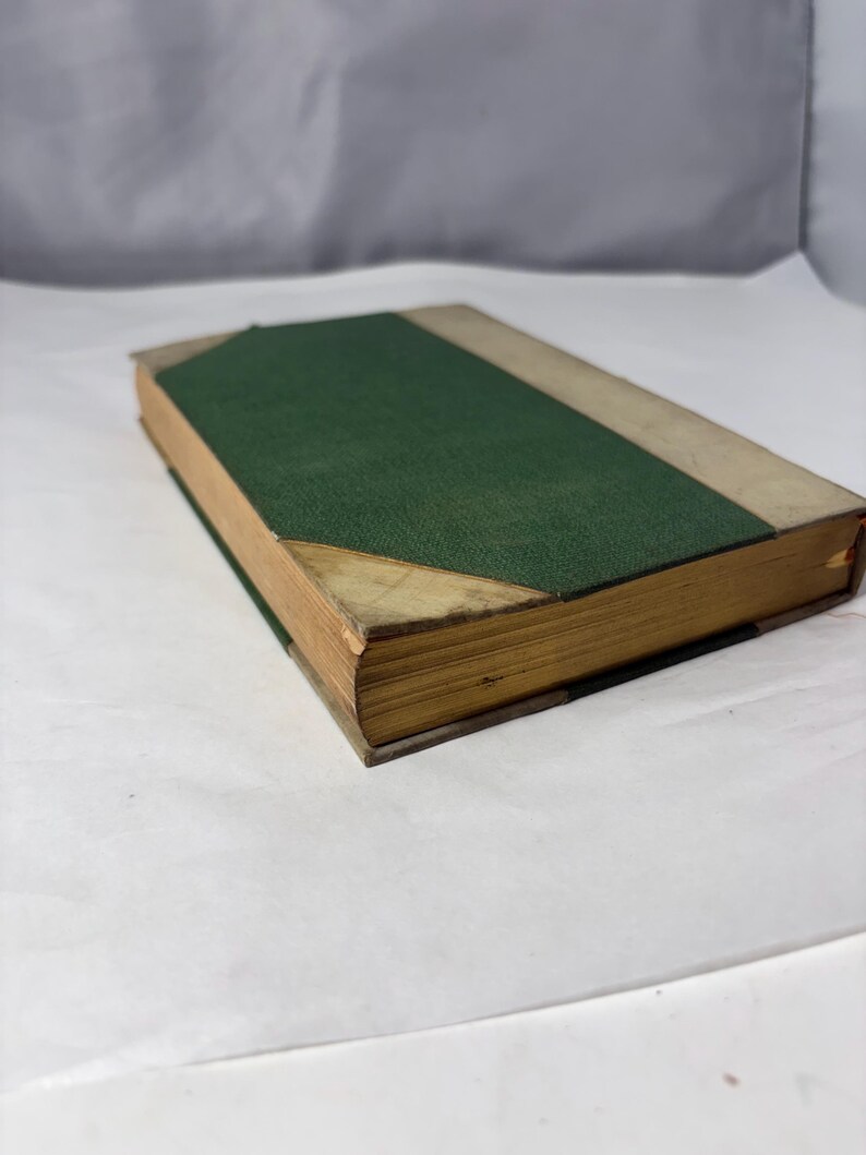Puede incluir: Un libro antiguo con una cubierta verde y bordes beige. El lomo del libro es verde y los bordes de las p&aacute;ginas est&aacute;n envejecidos. La cubierta tiene un aspecto texturizado y las esquinas muestran desgaste. El libro es probablemente una antig&uuml;edad o un objeto de colecci&oacute;n.