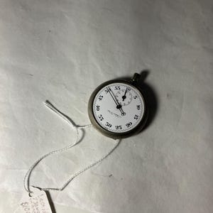 Könnte beinhalten: Eine silberfarbene Vintage-Stoppuhr mit weißem Zifferblatt und schwarzen Ziffern. Die Stoppuhr hat zwei Zifferblätter und eine kleine Krone. Ein weißes Band ist an der Stoppuhr befestigt mit einem Preisschild.