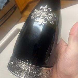 May include: A dark, glossy bottle with a silver decorative band at the base and a silver emblem near the top. The band features an engraved design with text. The bottle is held by a hand.