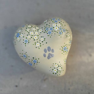 Könnte beinhalten: Ein weißer herzförmiger Keramik-Anhänger mit einem Pfotenabdruck und einem Muster aus blauen und weißen Blumen auf grünem Hintergrund.
