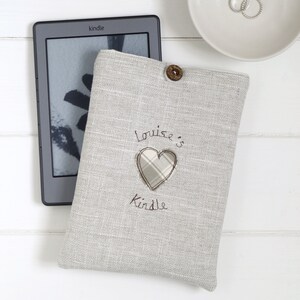 May include: A gray linen Kindle cover with a heart-shaped patch and the text "Louise's Kindle" embroidered on it.