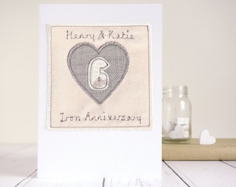 May include: A white card with a beige square on it. The square has a gray heart with a white number 6 inside. The text "Henry & Katie" and "Iron Anniversary" is embroidered on the beige square.