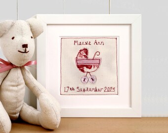 May include: A framed embroidered artwork featuring a baby carriage design in shades of pink and red. The artwork includes the name "Maeve Ann" and the date "17th September 2025". A stuffed teddy bear with a pink ribbon sits next to the frame.