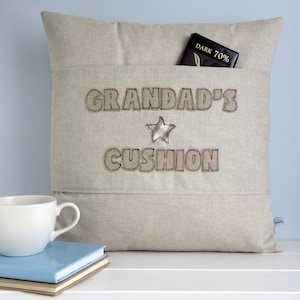 May include: A beige linen cushion with a pocket on the front. The cushion is embroidered with the text "Grandad's Cushion" and a star.