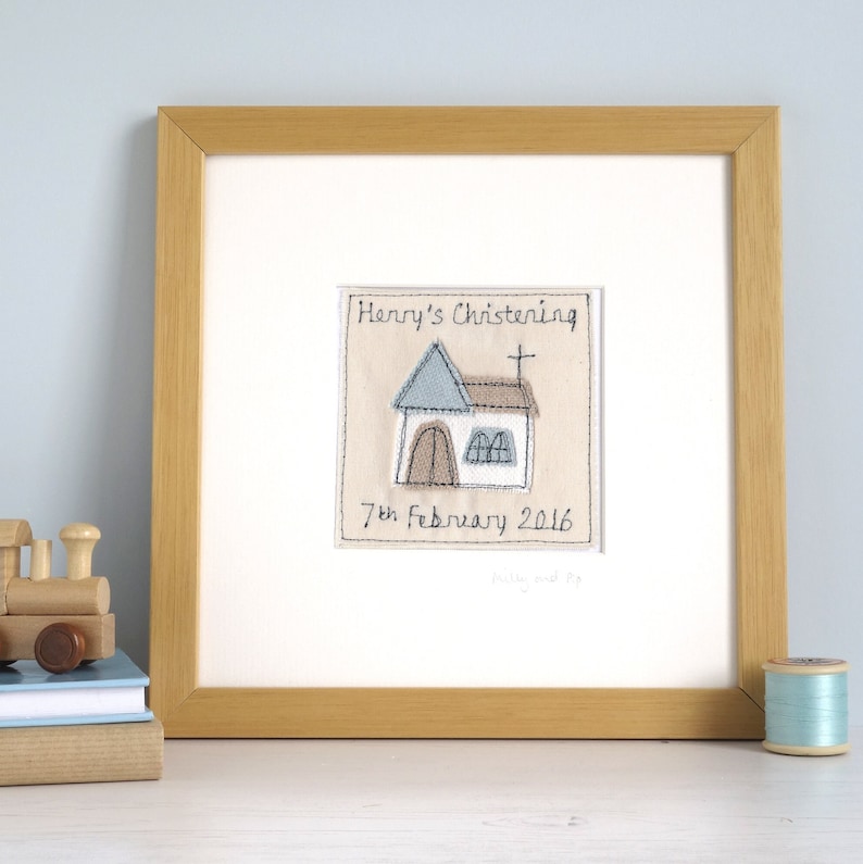 May include: A framed embroidered artwork featuring a church building with a cross on top, a blue roof, and a brown door. The text "Henry's Christening 7th February 2016" is embroidered below the church.