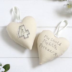 Può includere: Due ornamenti a forma di cuore in tessuto bianco. Un cuore ha il numero "4" ricamato su di esso. L'altro cuore ha il testo "My world My wife Forever x" ricamato su di esso.