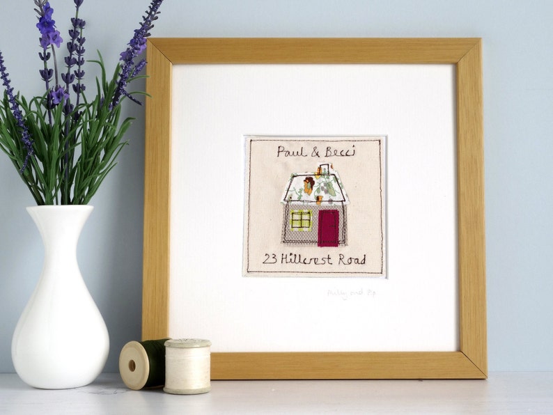 以下が含まれることがあります： 「Paul & Becci」と住所「23 Hillcrest Road」が書かれた、家の形をした刺繍が額装されています。