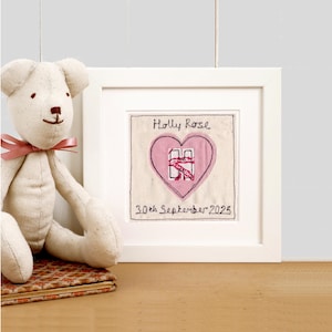 Puede incluir: Una obra de arte bordada enmarcada con el nombre "Holly Rose" y la fecha "30th September 2025" dentro de un corazón rosa. Un oso de peluche con un lazo rosa está junto al marco. El marco es blanco.
