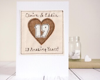 May include: A white greeting card with a fabric patch featuring a brown heart with the number 19 in white. The card reads "Claire & Eddie" and "19 Amazing Years!" in black script. A glass jar and heart-shaped decoration are in the background.
