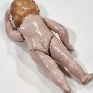 May include: A vintage porcelain doll with painted blonde hair and red eyes. The doll is wearing a pink dress and has a jointed body.