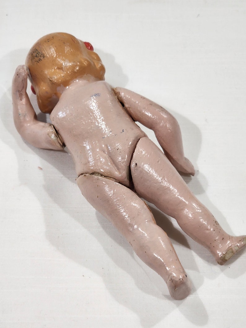 May include: A vintage porcelain doll with painted blonde hair and red eyes. The doll is wearing a pink dress and has a jointed body.