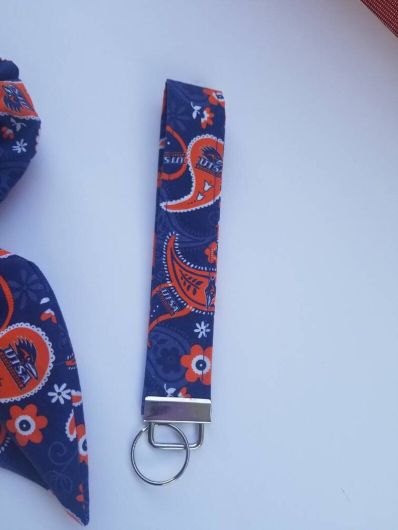May include: A blue and orange paisley patterned fabric keychain with a silver key ring. The fabric features the text "UTSA Roadrunners" repeated throughout the design.