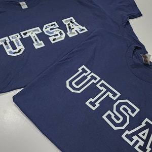 May include: Two navy blue t-shirts with the text "UTSA" printed on the front. The top shirt has the text in a camouflage pattern, while the bottom shirt has the text in white with a black outline.