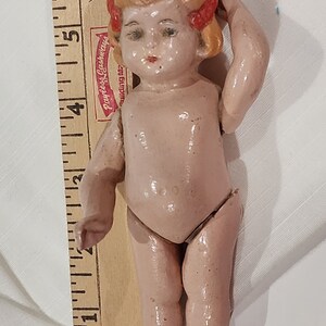 May include: A small, porcelain doll with a pink body, blonde hair, and red bows. The doll is standing with its right hand on its head and its left arm extended.