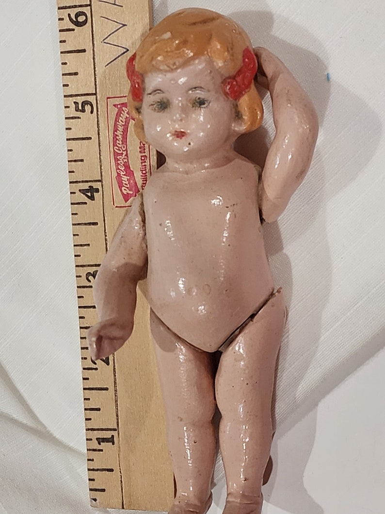 May include: A small, porcelain doll with a pink body, blonde hair, and red bows. The doll is standing with its right hand on its head and its left arm extended.