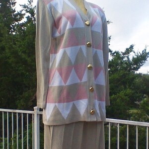 May include: A two-piece outfit on a mannequin. The top is a beige cardigan with a diamond pattern in pink, white, and beige. It has gold buttons. The skirt is a matching beige color and pleated.
