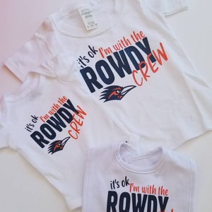 May include: A set of three white t-shirts with a blue and orange graphic design. The graphic features a bird with the text "it's ok I'm with the ROWDY CREW". The shirts are in different sizes, with one being a baby bib.