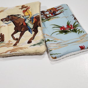 May include: Two fabric pot holders with a cowboy theme. One is beige with a cowboy riding a horse and the other is blue with a floral pattern.
