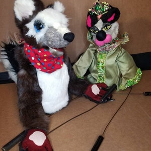 May include: Two hand puppets, one with a black and white fur design and a red and white polka dot scarf, the other with a green and gold costume and a crown. Both puppets have black eyes and pink noses.