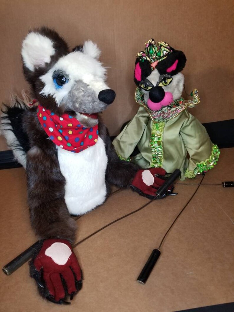 May include: Two hand puppets, one with a black and white fur design and a red and white polka dot scarf, the other with a green and gold costume and a crown. Both puppets have black eyes and pink noses.
