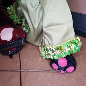 May include: A green and gold puppet with a black and pink foot. The puppet is wearing a green and gold costume with sequins. The puppet is sitting on a brown surface.